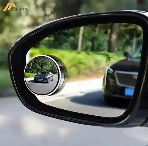 Car side mirror with a small round mirror attached, showing a blurred background of trees and road.