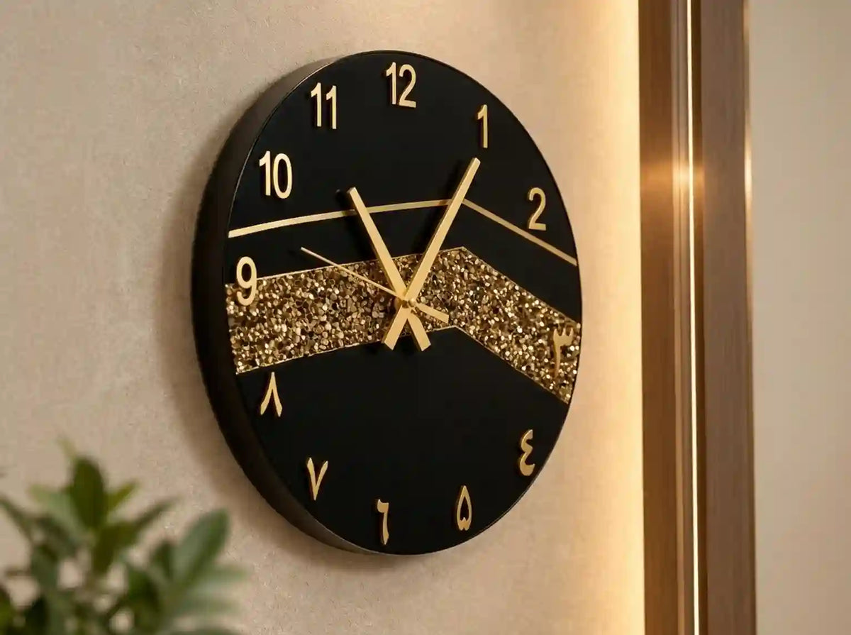 A round black wall clock with gold Arabic numerals, gold hands, and a glittery gold chevron accent across the center, mounted on a beige wall.