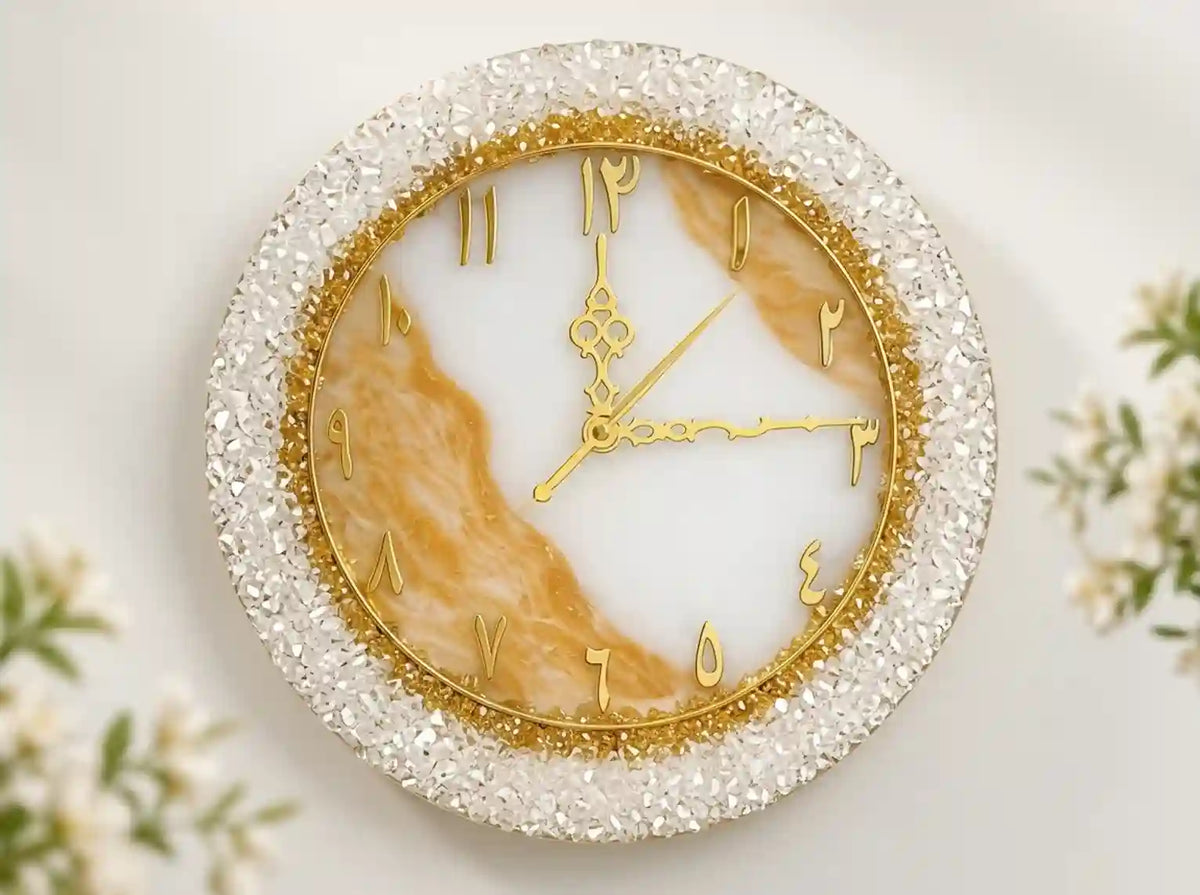 A round wall clock with a white and gold marbled face, gold Arabic numeral hour hands, and a sparkling clear rhinestone outer bezel, shown against a light background with soft floral props.