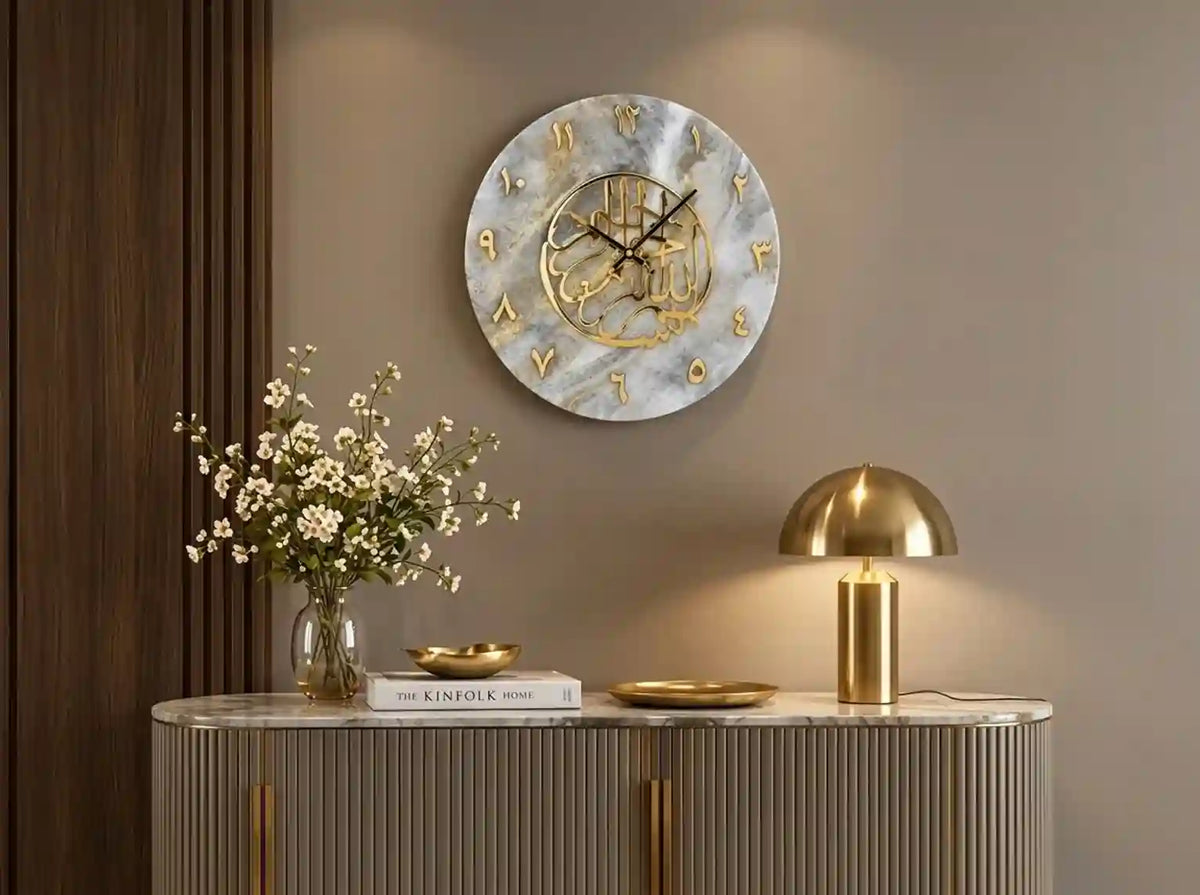 A round wall clock with a white marble pattern face and gold Arabic numerals, styled in a modern home interior above a console table with a gold lamp and flowers.