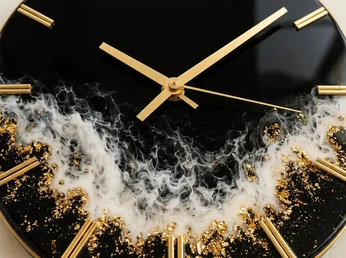 A close-up of a round wall clock with a glossy black face, gold hands, gold linear hour markers, and a white marbled pattern with scattered gold leaf flakes around the lower half.