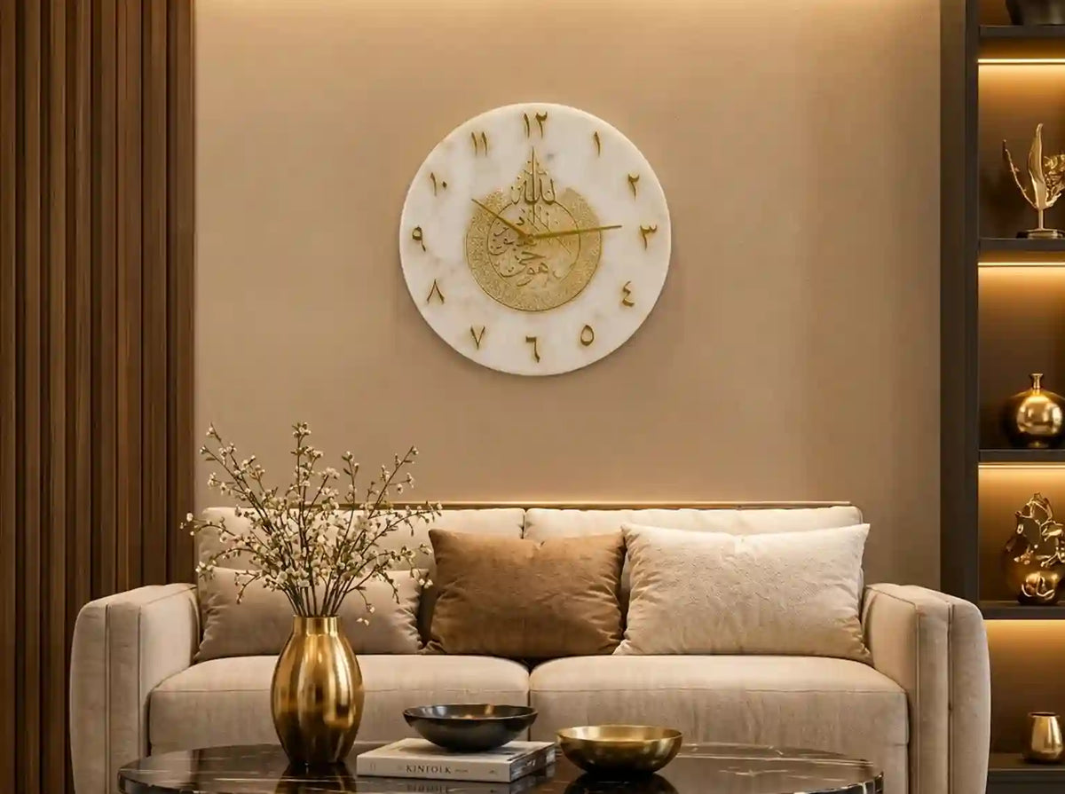 A round white marble wall clock with gold Roman numerals and a gold central emblem, mounted on a beige wall above a modern beige sofa in a styled living room.