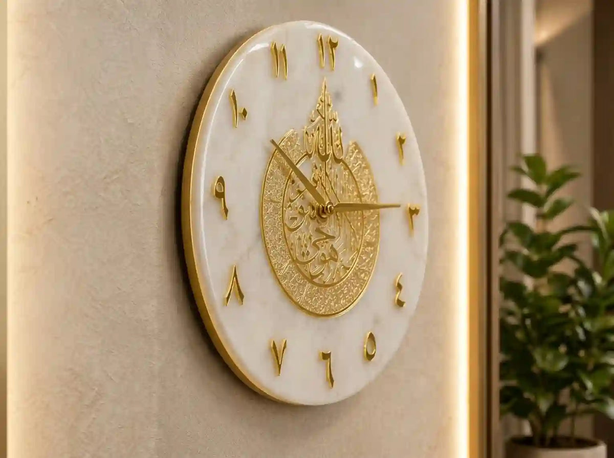 An oval wall clock with a white marble-look face and ornate gold details, mounted on a beige wall with a softly lit interior and a plant visible in the background.