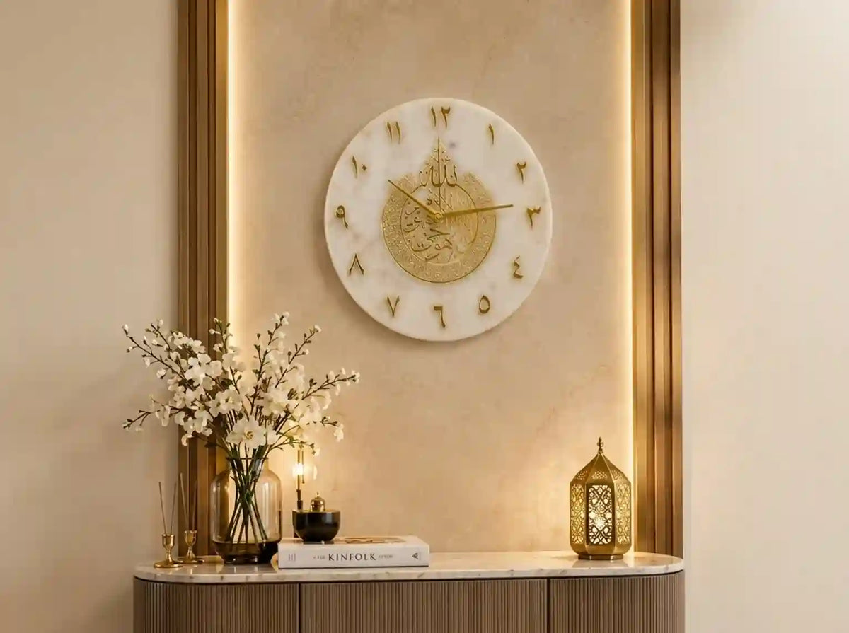 A round white marble wall clock with gold Roman numerals and gold hands, mounted on a beige wall with warm backlighting. Below the clock, a console table holds a vase with white blossoms, a candle, a book, and a gold lantern.