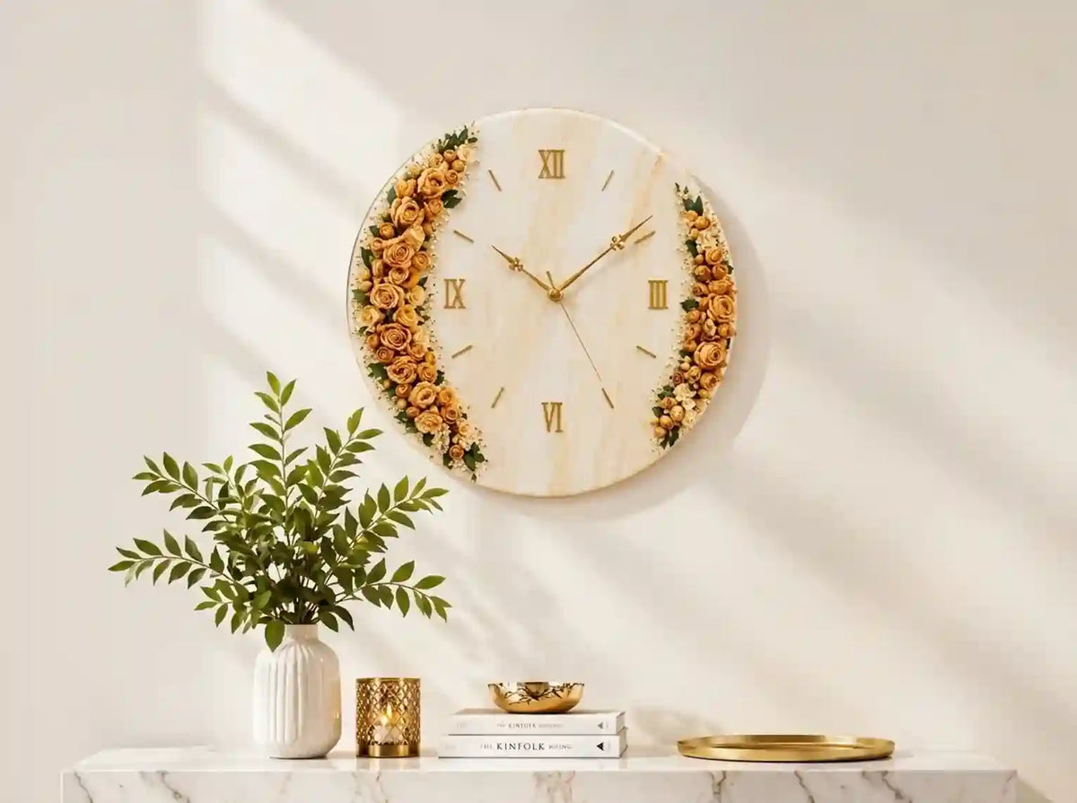A round wall clock with a cream marble-like face, gold Roman numerals, and gold hands, decorated with dried floral garlands on the left and right sides. The clock is mounted on a warm beige wall with soft window shadows, above a marble console table styled with a green plant in a white vase, a gold candle holder, books, and a gold tray.