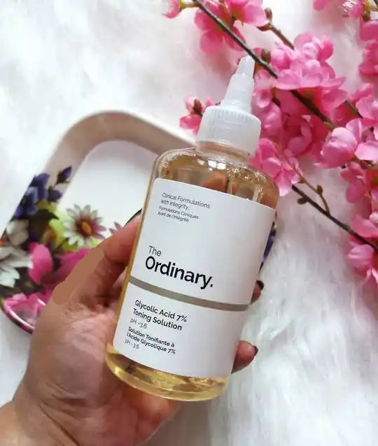 Bottle of 'The Ordinary' skincare solution held by a hand with pink flowers in the background