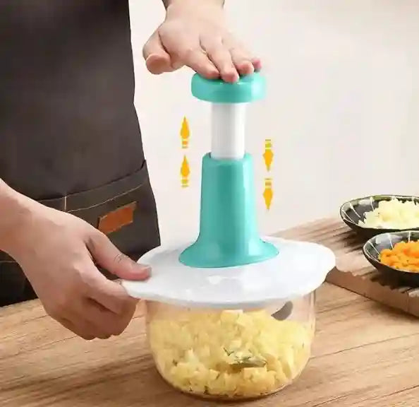 Person using a manual food chopper with a clear container filled with chopped vegetables on a wooden surface.