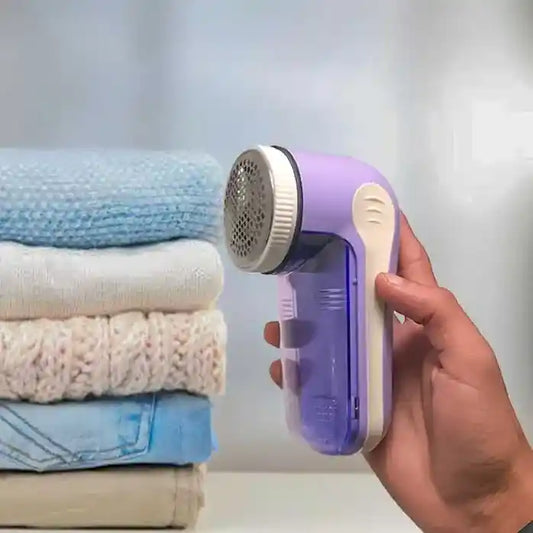 Hand holding a purple fabric shaver with folded clothes in the background