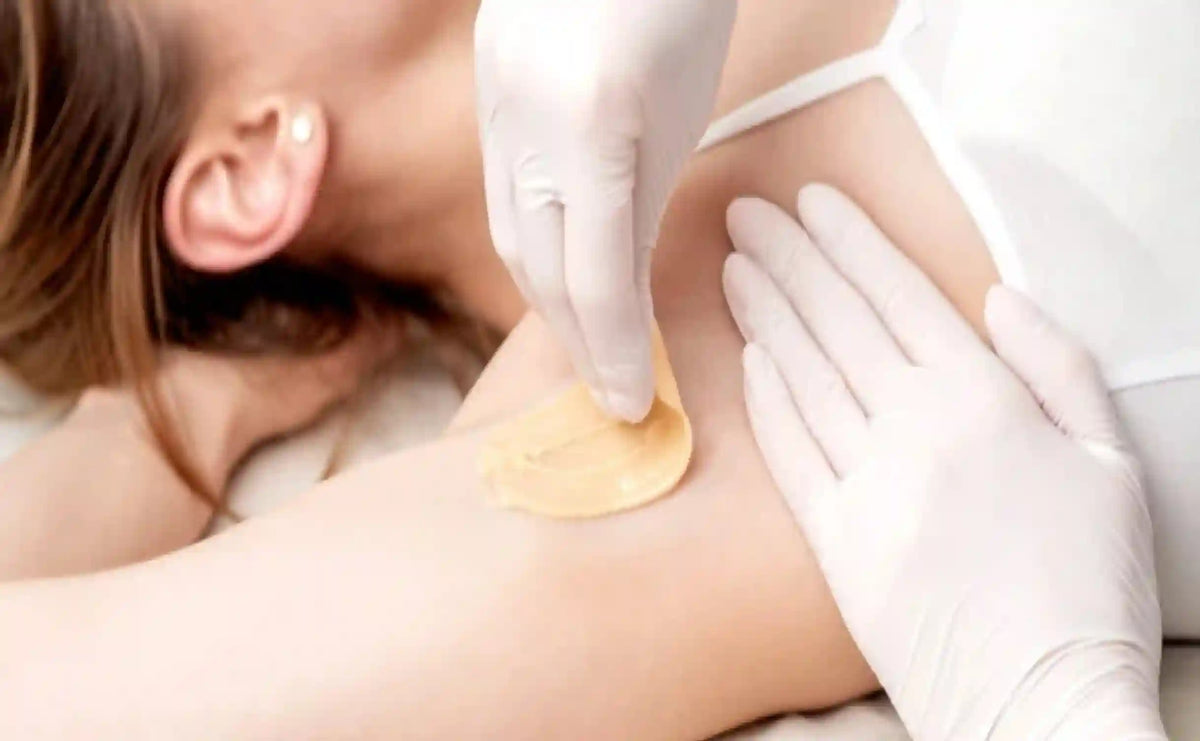 A close-up of a person lying down while another person wearing white gloves applies a beige hair removal patch to the upper arm.