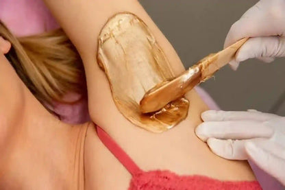 A close-up of a person's underarm with a thick, glossy golden-brown hair removal wax or depilatory cream being spread with a wooden spatula by a gloved hand.