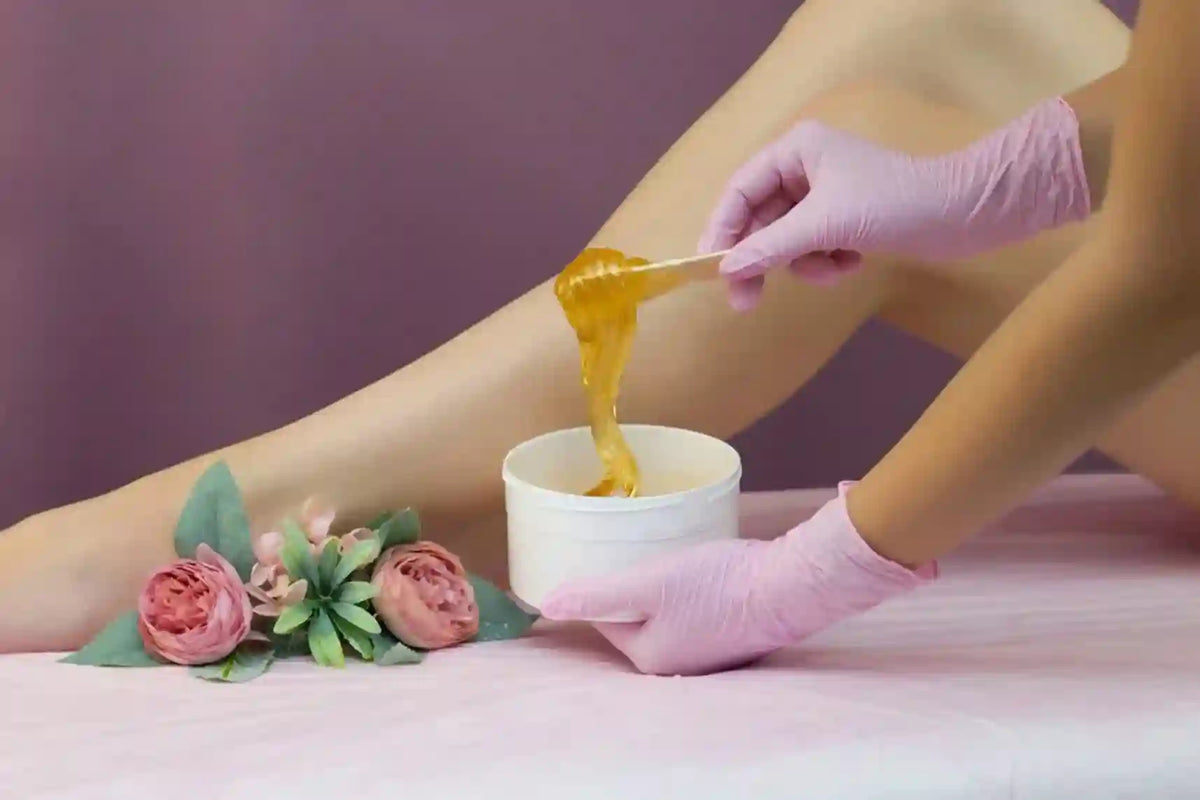 A close-up of a person's leg with pink gloves holding a small white cup while applying a thick, golden honey-like substance from a wooden dipper. Pink roses are placed on the pink surface beside the leg, against a soft mauve background.