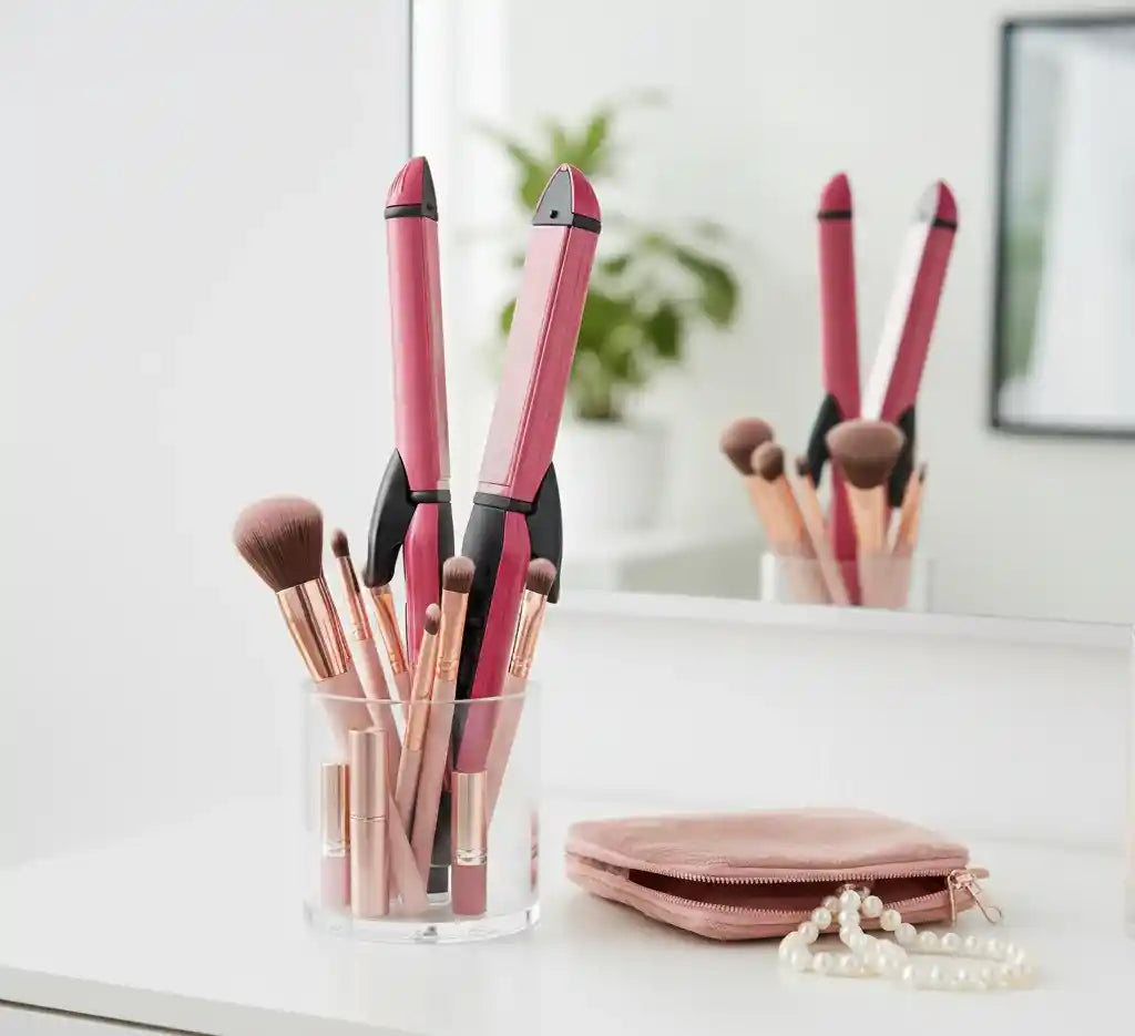 Makeup brushes and a hair straightener in a glass holder on a white surface.