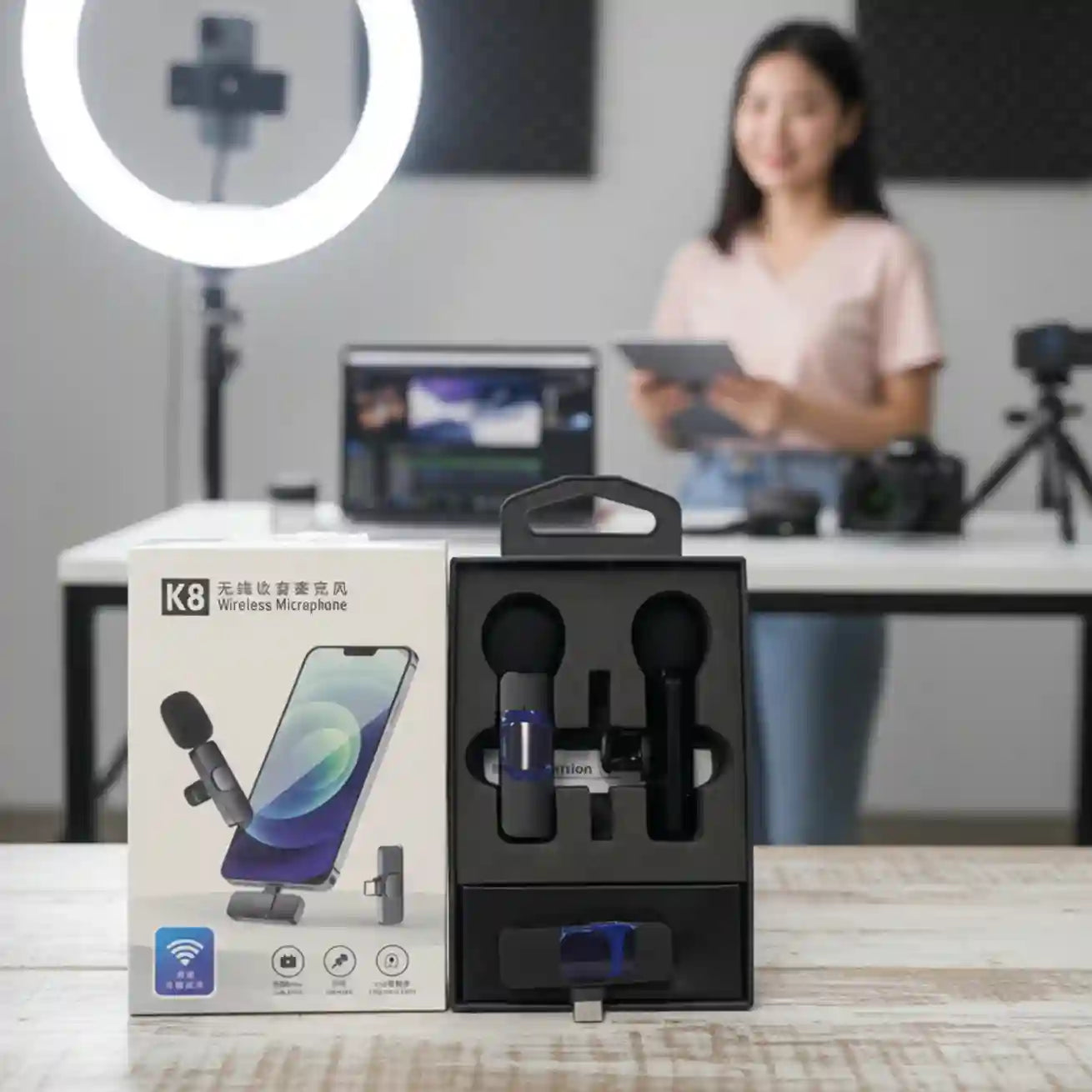 K8 wireless microphone set with packaging on a desk, blurred background of a person using a tablet.