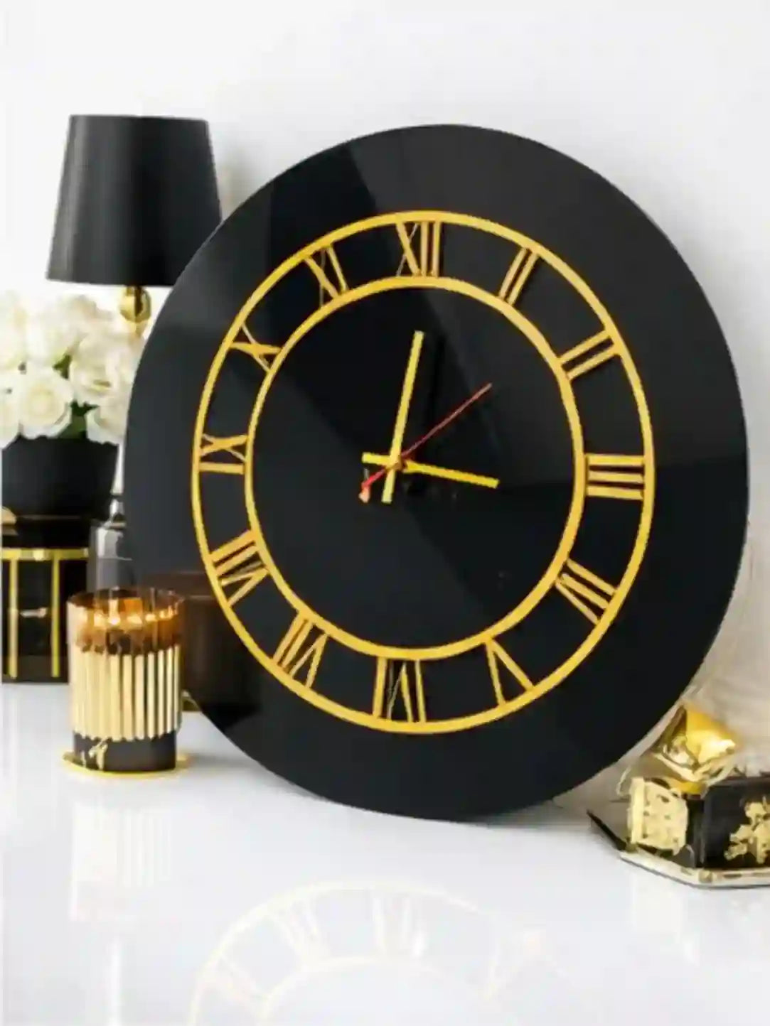 Black wall clock with gold Roman numerals on a white surface with decorative items.