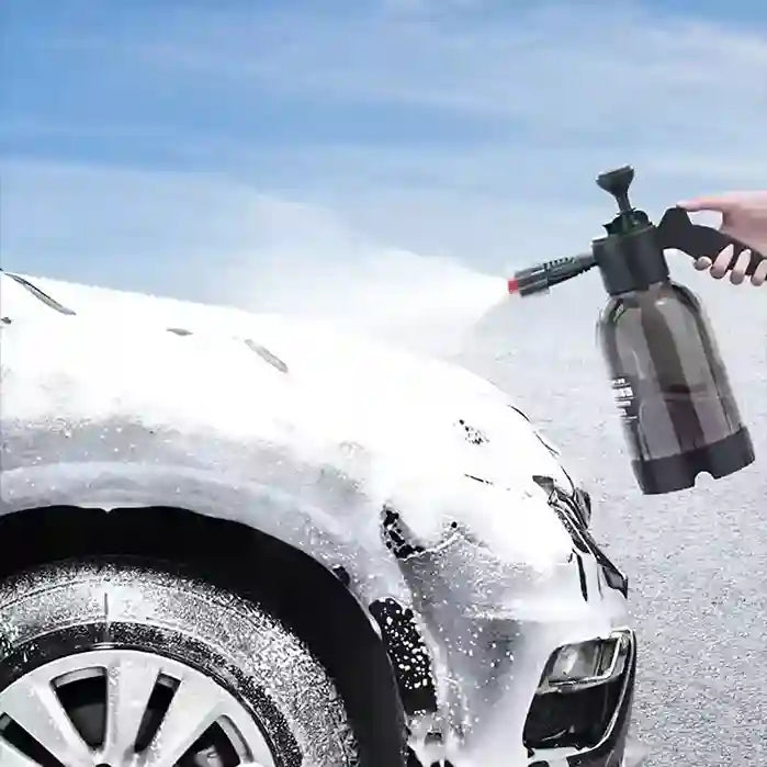 Person using a spray bottle to wash a car with soap suds on a clear day.
