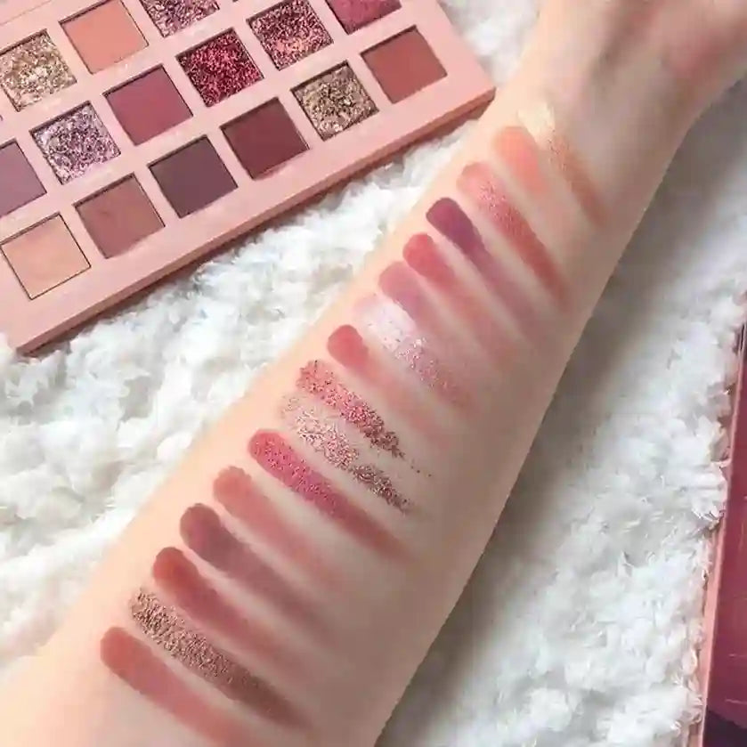 Makeup swatches on a person's arm with a pink eyeshadow palette in the background