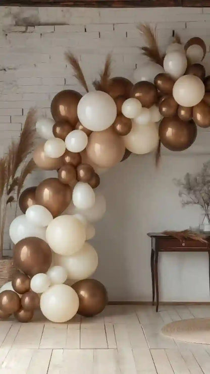 Balloon arch with gold, white, and brown balloons against a white wall.