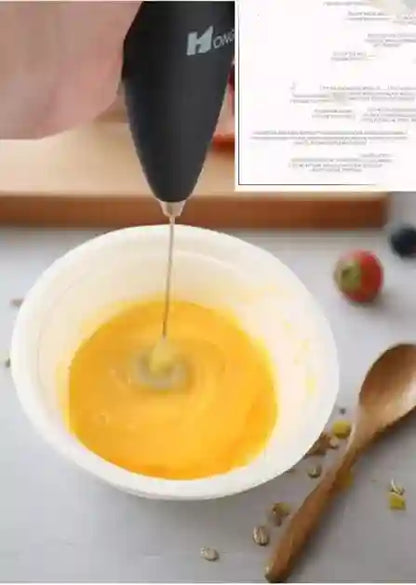 Handheld mixer being used to whip eggs in a bowl with a wooden spoon and ingredients in the background.