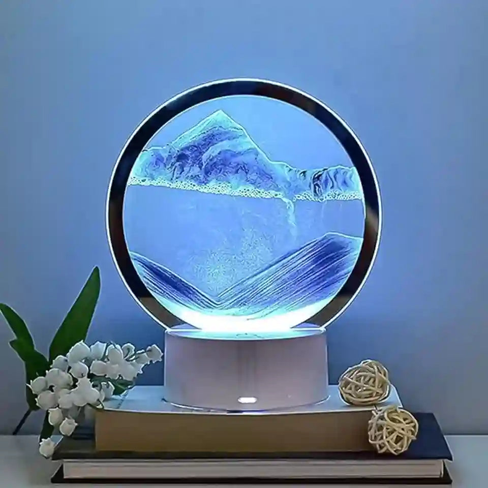A circular LED sand art lamp displaying a mountain landscape inside a clear glass dome, resting on a white cylindrical base atop stacked books with decorative flowers and woven balls against a blue wall.