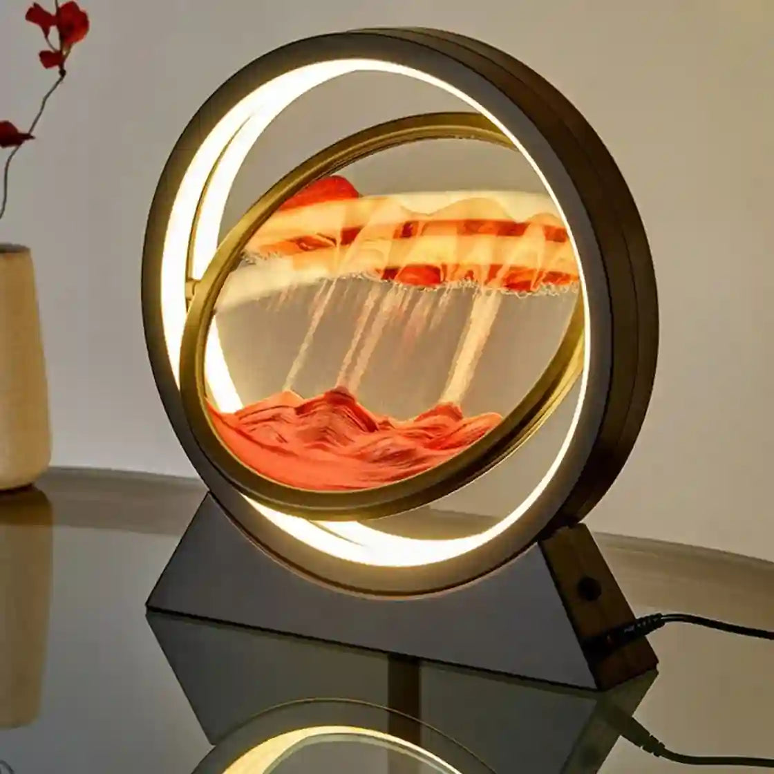A circular LED-lit sand art display with flowing orange and red sand inside a transparent dome, mounted on a dark rectangular base on a reflective tabletop.