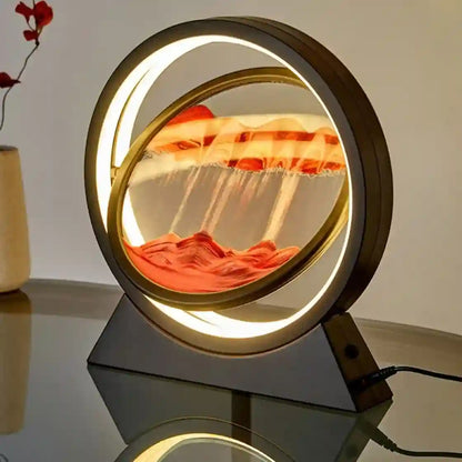 A circular LED-lit sand art display with flowing orange and red sand inside a transparent dome, mounted on a dark rectangular base on a reflective tabletop.