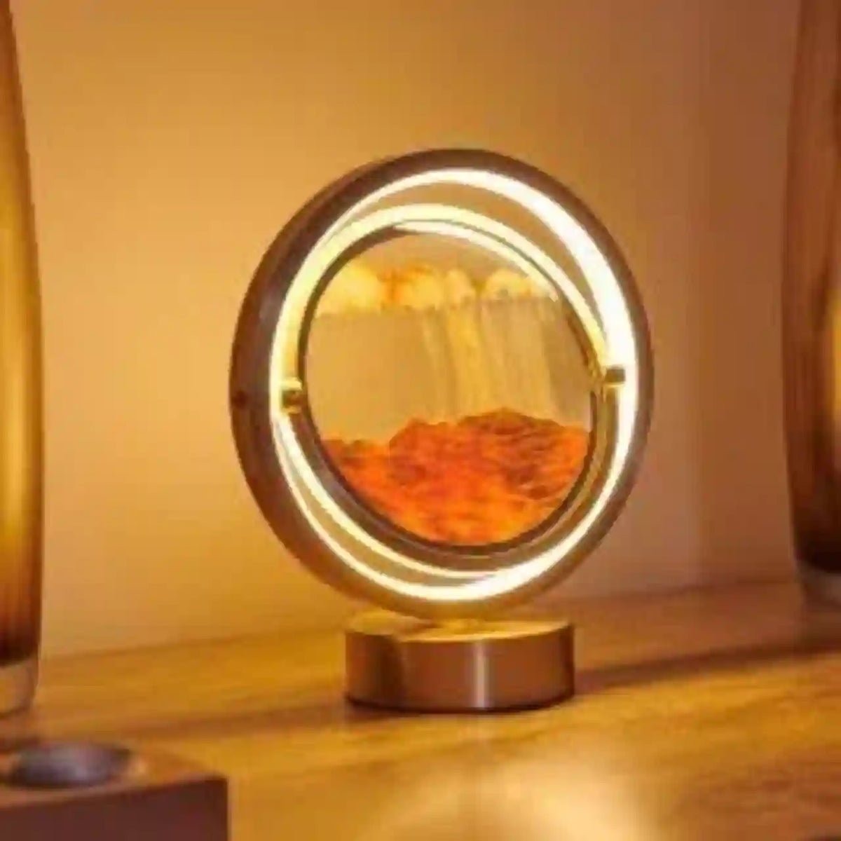 A modern oval LED night light with a gold-toned metal frame and base, featuring a glowing inner ring and a reflective center showing warm orange and yellow light patterns, set on a wooden surface.