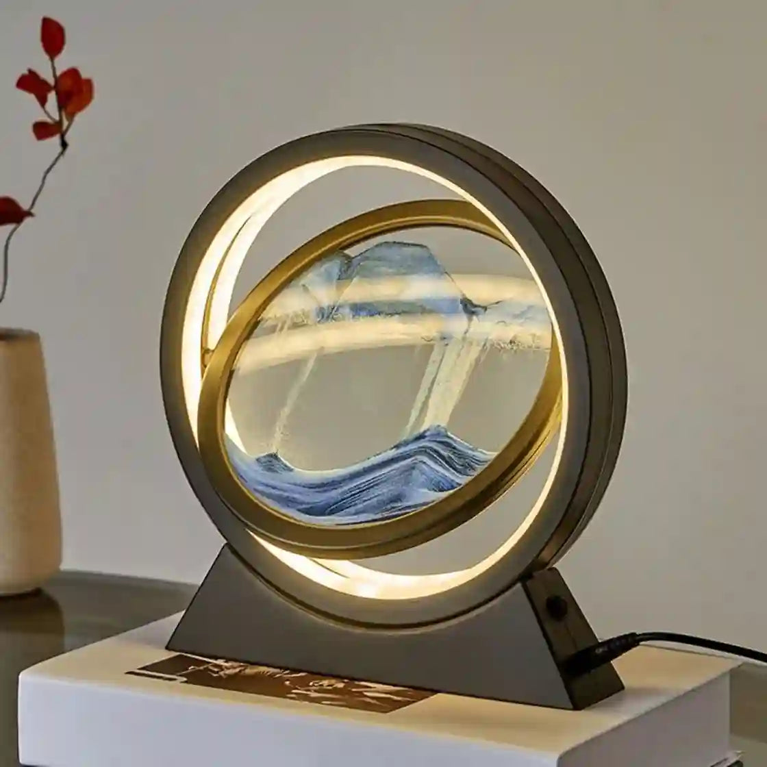 A modern circular LED desk lamp with a gold-toned frame and a swirling sand or glass interior showing a mountain-and-water scene, placed on a white book with a vase in the background.