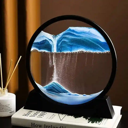 A circular sand art lamp with a black frame and stand, showing blue and white flowing sand patterns resembling water or waves. It is styled on top of books next to a reed diffuser against a warm brown background.
