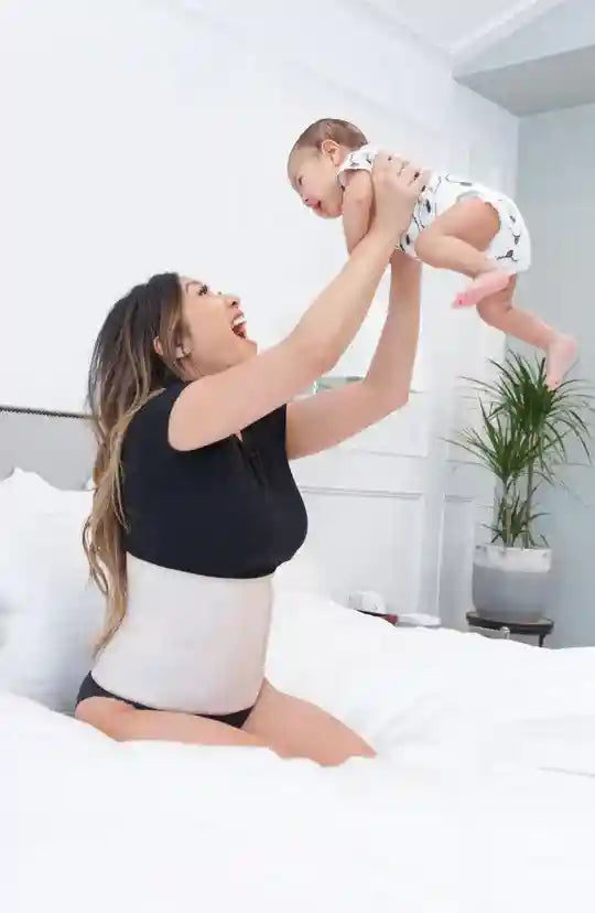 Woman lifting a baby in a room with a plant and bed.