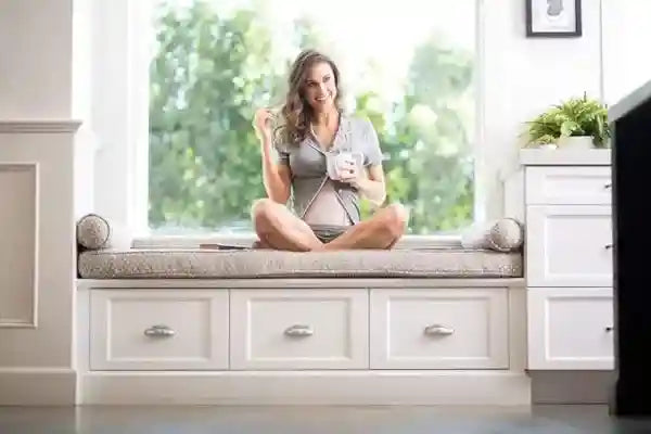 Woman sitting on a window seat with a cup, surrounded by white furniture and greenery