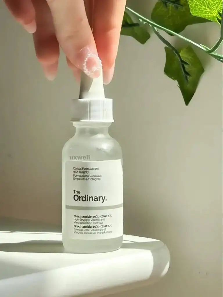 Bottle of 'The Ordinary' serum with a dropper, held by a hand with green leaves in the background.