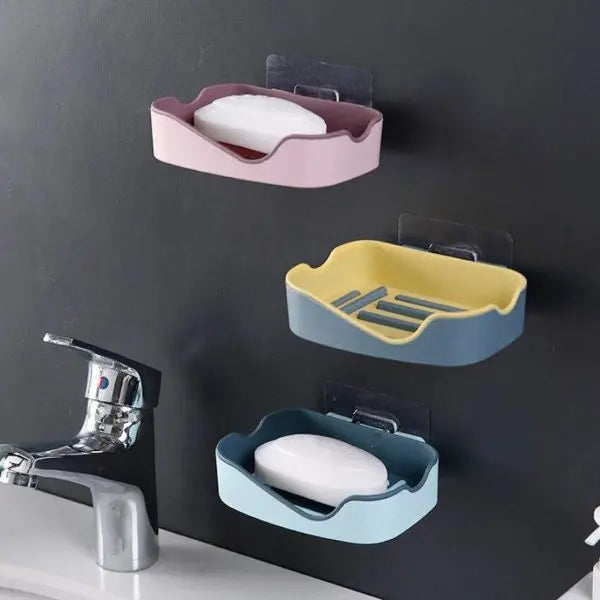 Three wall-mounted soap holders in pink, yellow, and blue on a black wall with a faucet in the foreground.