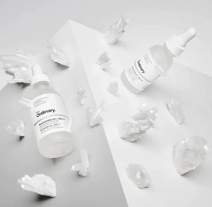 Two 'The Ordinary' skincare bottles on a light gray background with ice cubes.