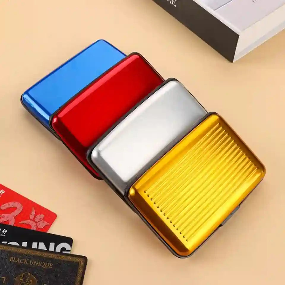 Four slim rectangular metal card cases in blue, red, silver, and gold, arranged diagonally on a beige surface with a few cards and a book corner visible as props.