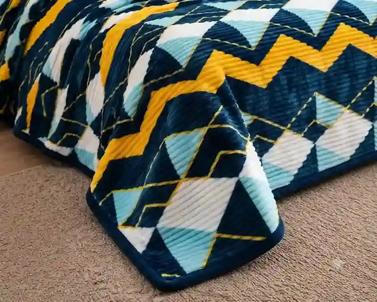 A close-up of a plush blanket with a geometric zigzag and diamond pattern in navy blue, yellow, light blue, and white, draped over a bed on a beige carpet.