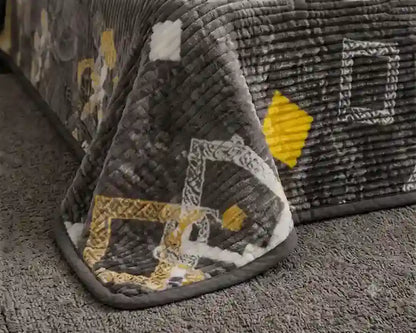 A close-up of a textured gray quilted blanket draped over the corner of a bed, featuring a geometric pattern with yellow and white accents on a carpeted floor.