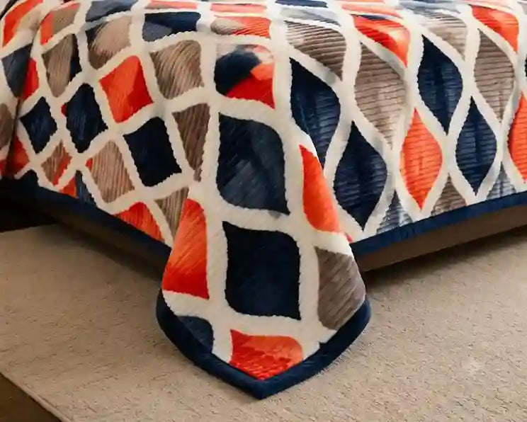 A close-up of a textured blanket draped over the corner of a bed, featuring a bold geometric diamond pattern in orange, navy blue, gray, and white.