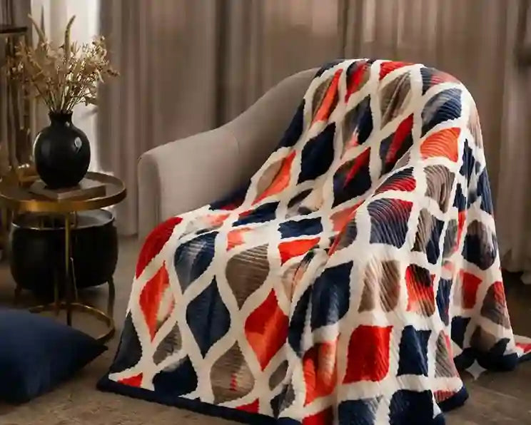 A textured throw blanket draped over a light gray armchair in a cozy living room setting. The blanket features a bold geometric diamond pattern in navy blue, red-orange, and cream white.