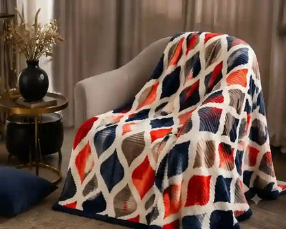 A textured throw blanket draped over a light gray armchair in a cozy living room setting. The blanket features a bold geometric diamond pattern in navy blue, red-orange, and cream white.