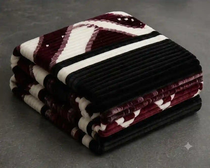 A stack of folded plush corduroy blankets in a color-blocked pattern of burgundy, cream, and black, set against a dark gray textured background.