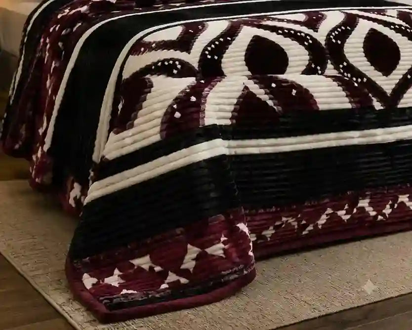 A close-up of a quilted blanket draped over a bed, featuring a geometric pattern in deep burgundy, black, and cream.