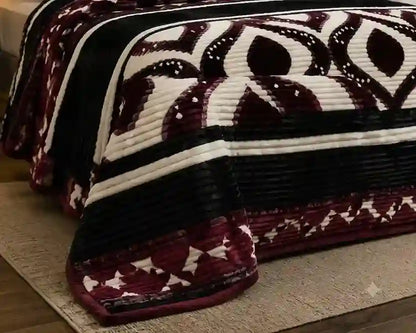 A close-up of a quilted blanket draped over a bed, featuring a geometric pattern in deep burgundy, black, and cream.