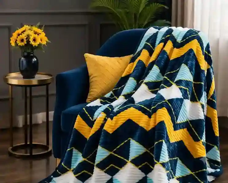 A plush throw blanket with a geometric chevron and diamond pattern in navy blue, yellow, light blue, and white, draped over a dark blue armchair in a styled living room setting.