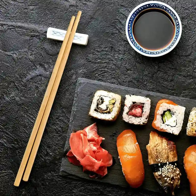 Assorted sushi on a black slate board with chopsticks and a bowl of soy sauce on a dark surface.