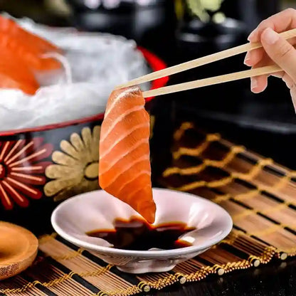 Person using chopsticks to dip salmon sashimi into soy sauce on a wooden mat.