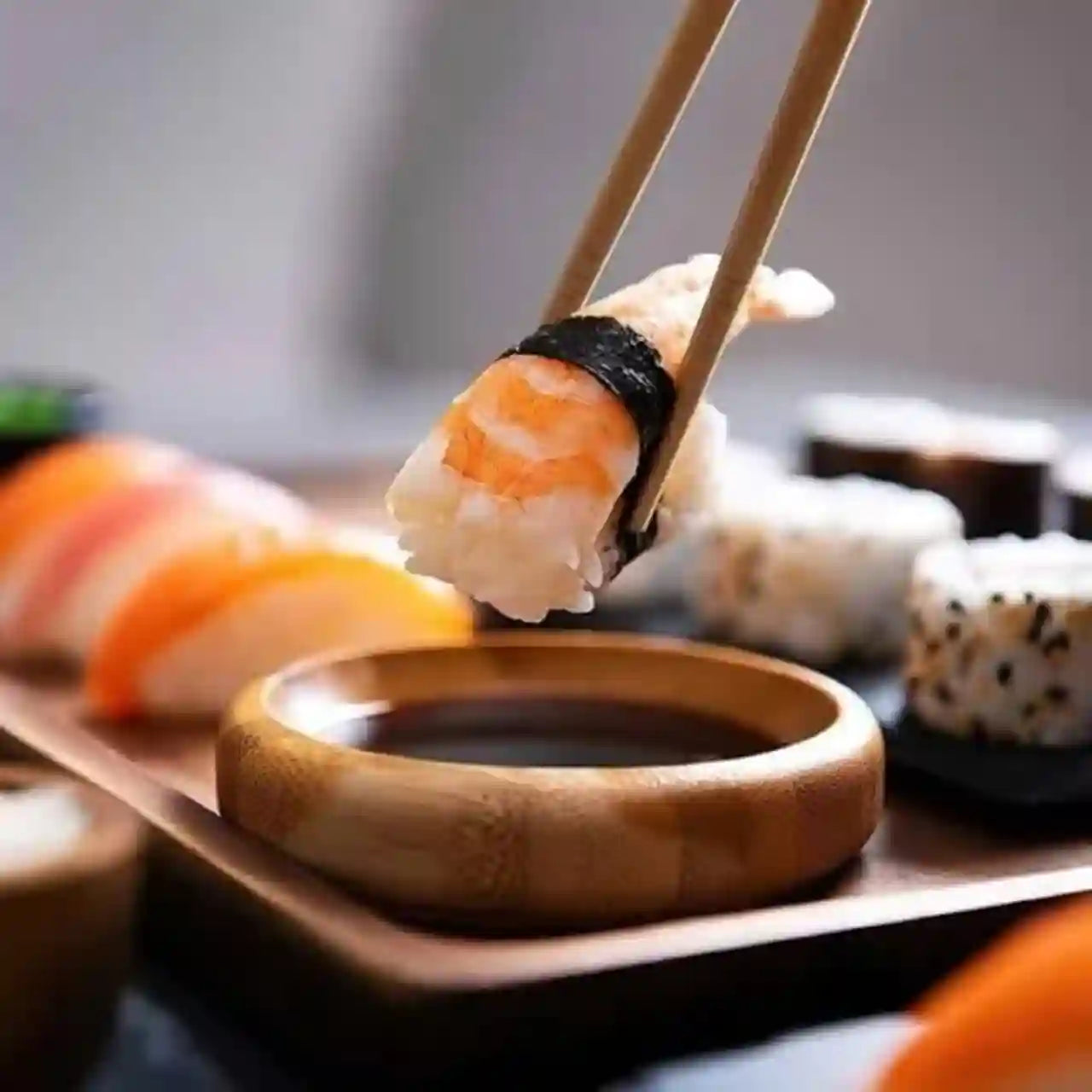 A close-up shot of a piece of sushi held by wooden chopsticks above a small wooden dipping bowl, with other sushi pieces blurred in the background.