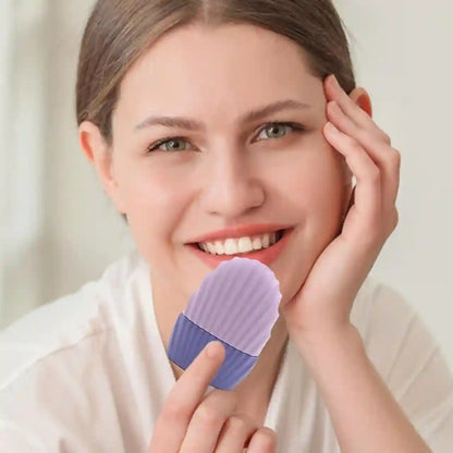 A smiling woman with her face partially obscured by a lavender and purple silicone beauty sponge held near her cheek, wearing a white top against a soft light background.