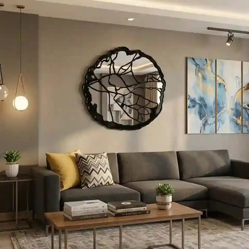 A modern living room featuring a large, organic-shaped black metal wall mirror with a leaf-like pattern above a gray sectional sofa. The room has a warm neutral color palette with a gray wall, a wooden coffee table, and abstract blue and gold wall art.