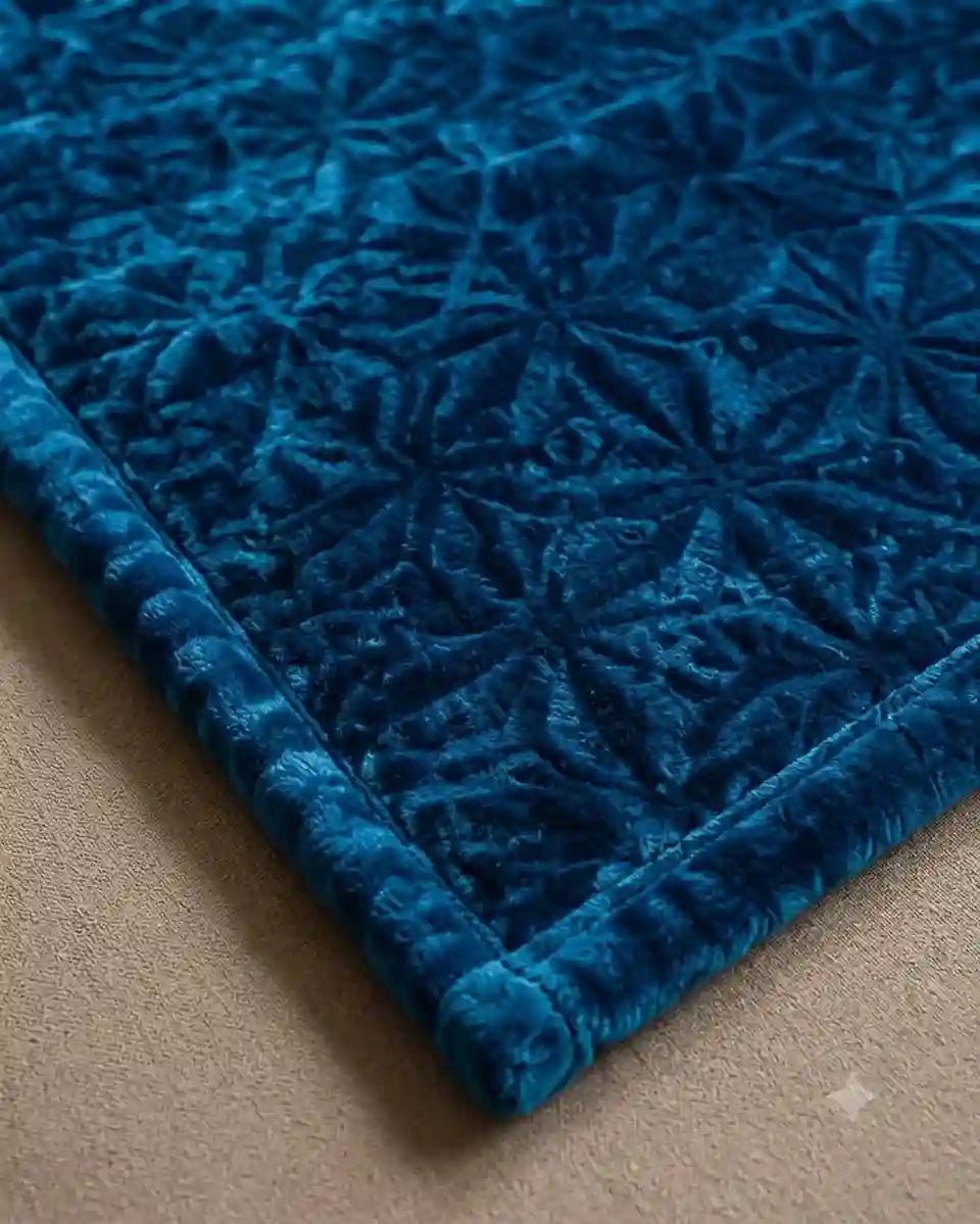 A close-up of a plush velvet textile, likely a blanket or throw, featuring a starburst quilted pattern and a thick ribbed border, shown on a neutral beige surface.
