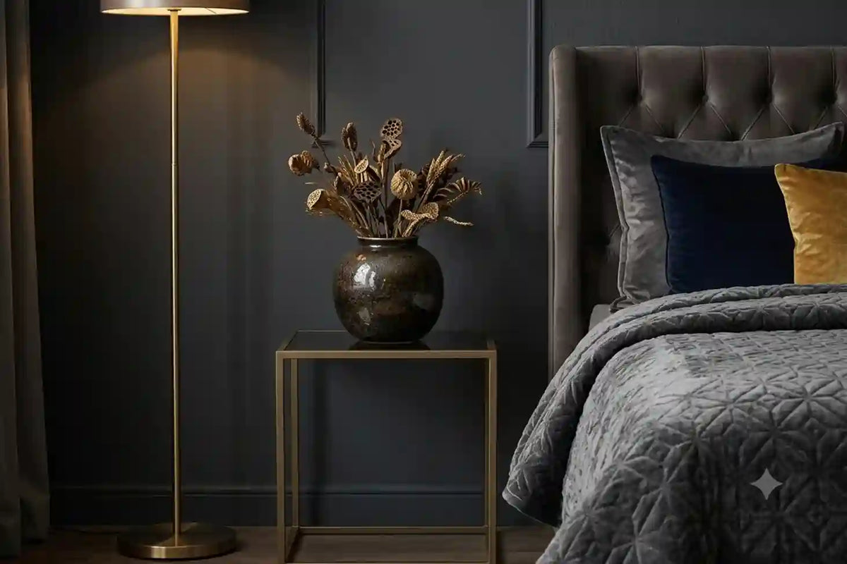 A styled bedroom corner featuring a dark gray tufted headboard, gray quilted bedding, a dark blue and yellow accent pillow, a gold floor lamp, and a small gold side table holding a dark vase with dried botanicals.